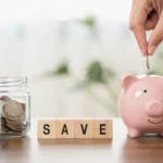 Hand putting a coin into a pink piggy bank beside a jar of savings