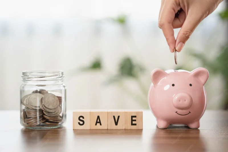 Hand putting a coin into a pink piggy bank beside a jar of savings